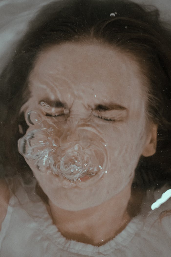 From above of female with closed eyes immersed in water in bathtub forming bubbles on surface while holding breath at home